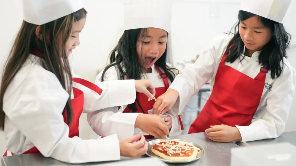 Three kids in chef hats and red aprons topping a mini pizza